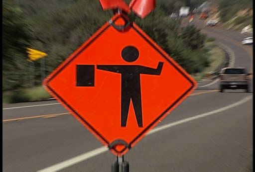 Safety orange diamond sign with figure of flagger beside roadway