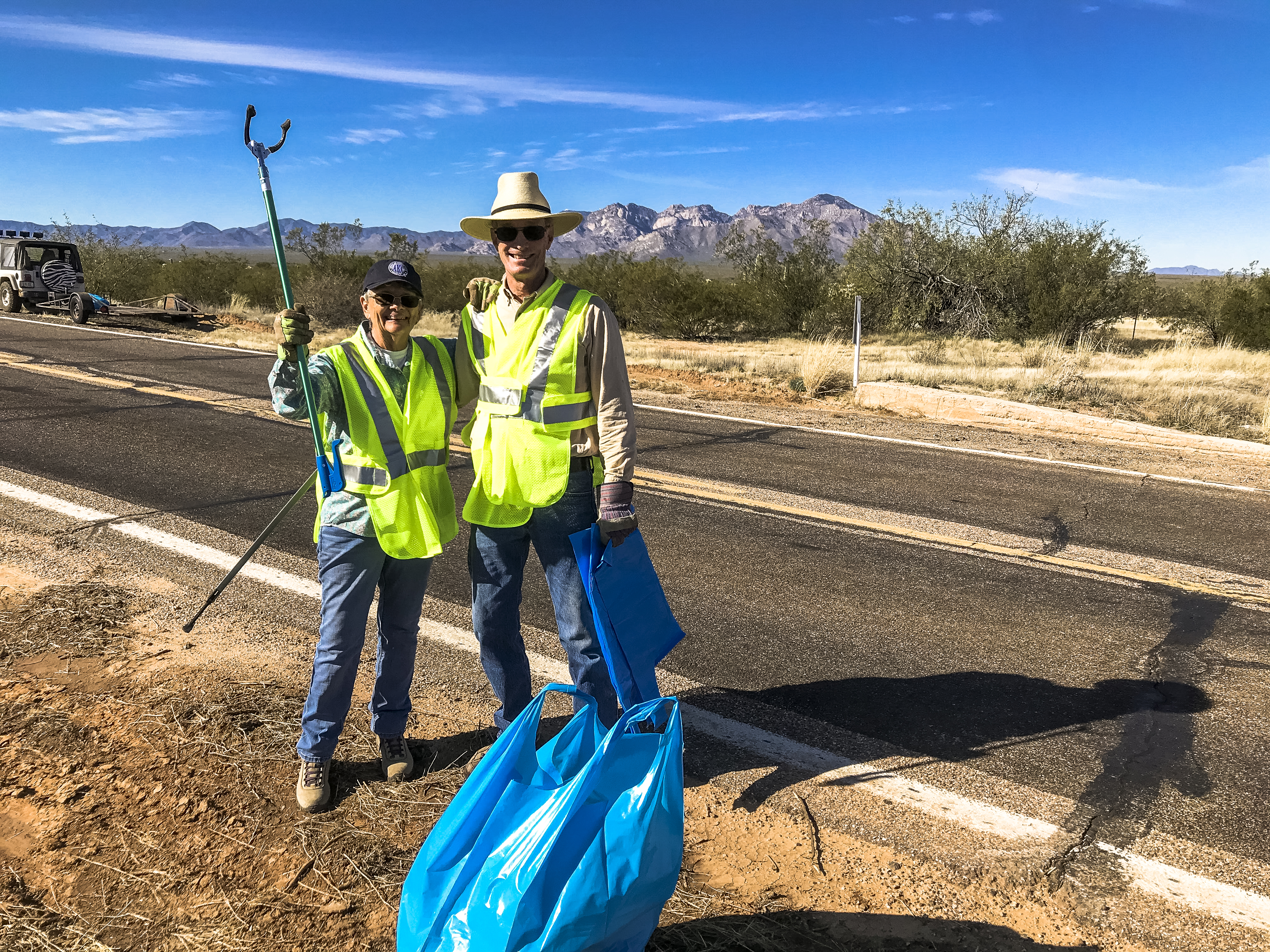 Two people in yellow vests standing on a rural roadside with blue trashbags at their feet.