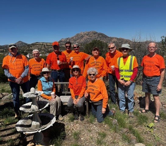 A group of 12 people, all wearing traffic safety orange shirts and members of a volunteer litter-pick-up group, gather for a group photo with a mountain in the background.