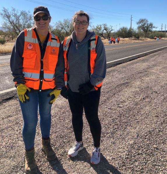 Two people stand on a highway shoulder in orange safety vests. 