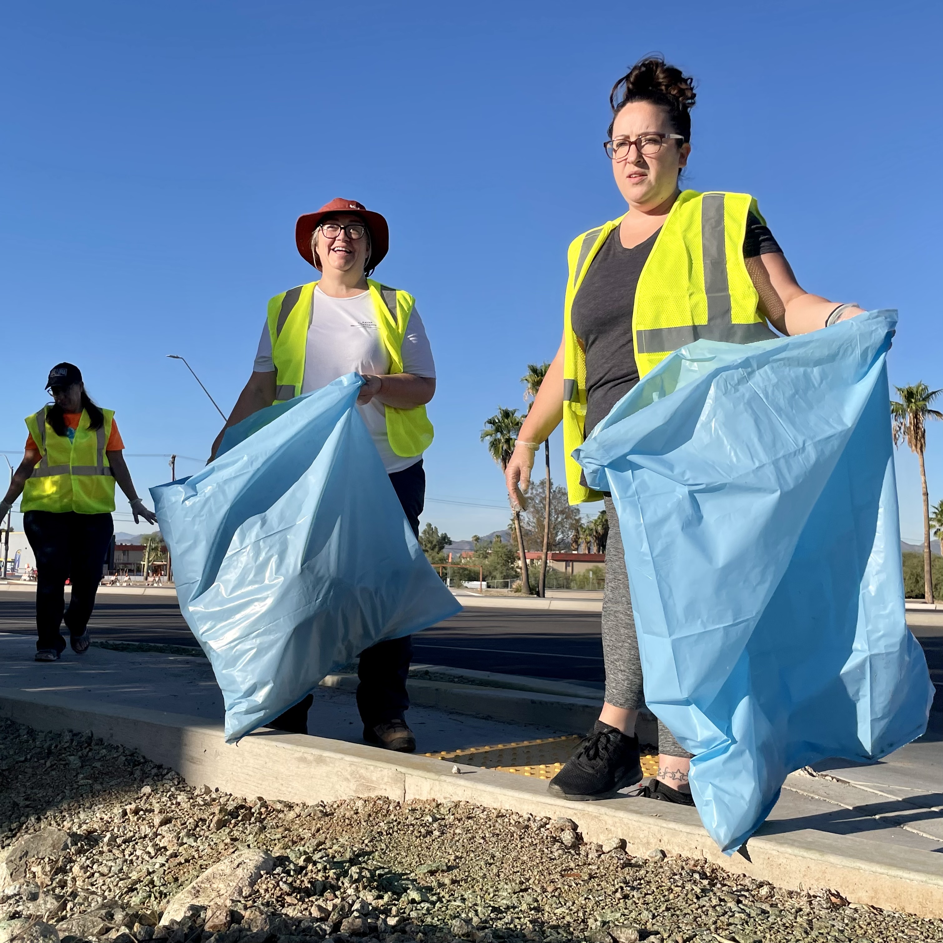 Three people in bright yellow reflective safety vests pick up litter on the side of a highway.