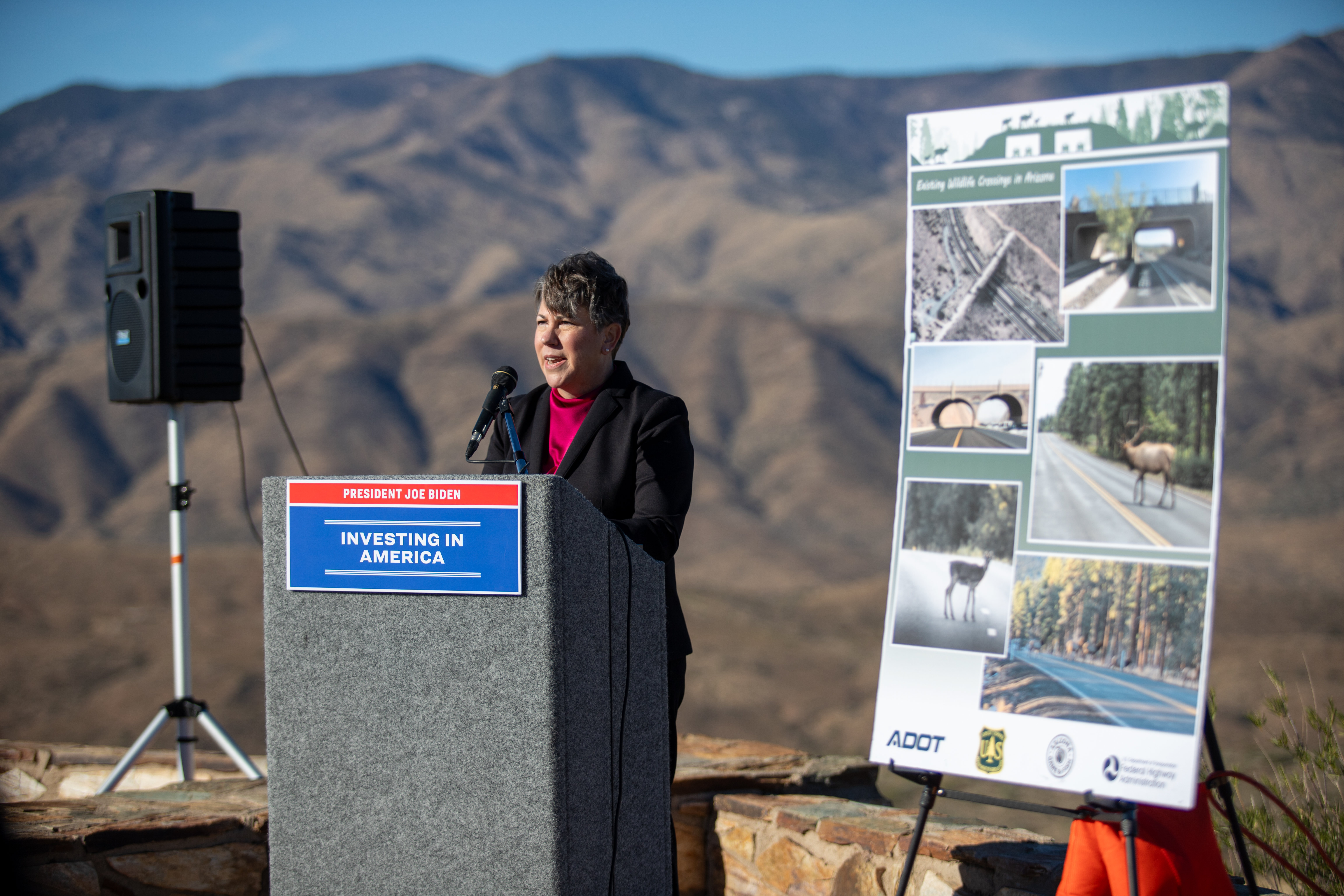 A woman stands at a podium at an outdoors press conference. Mountains are in the background.