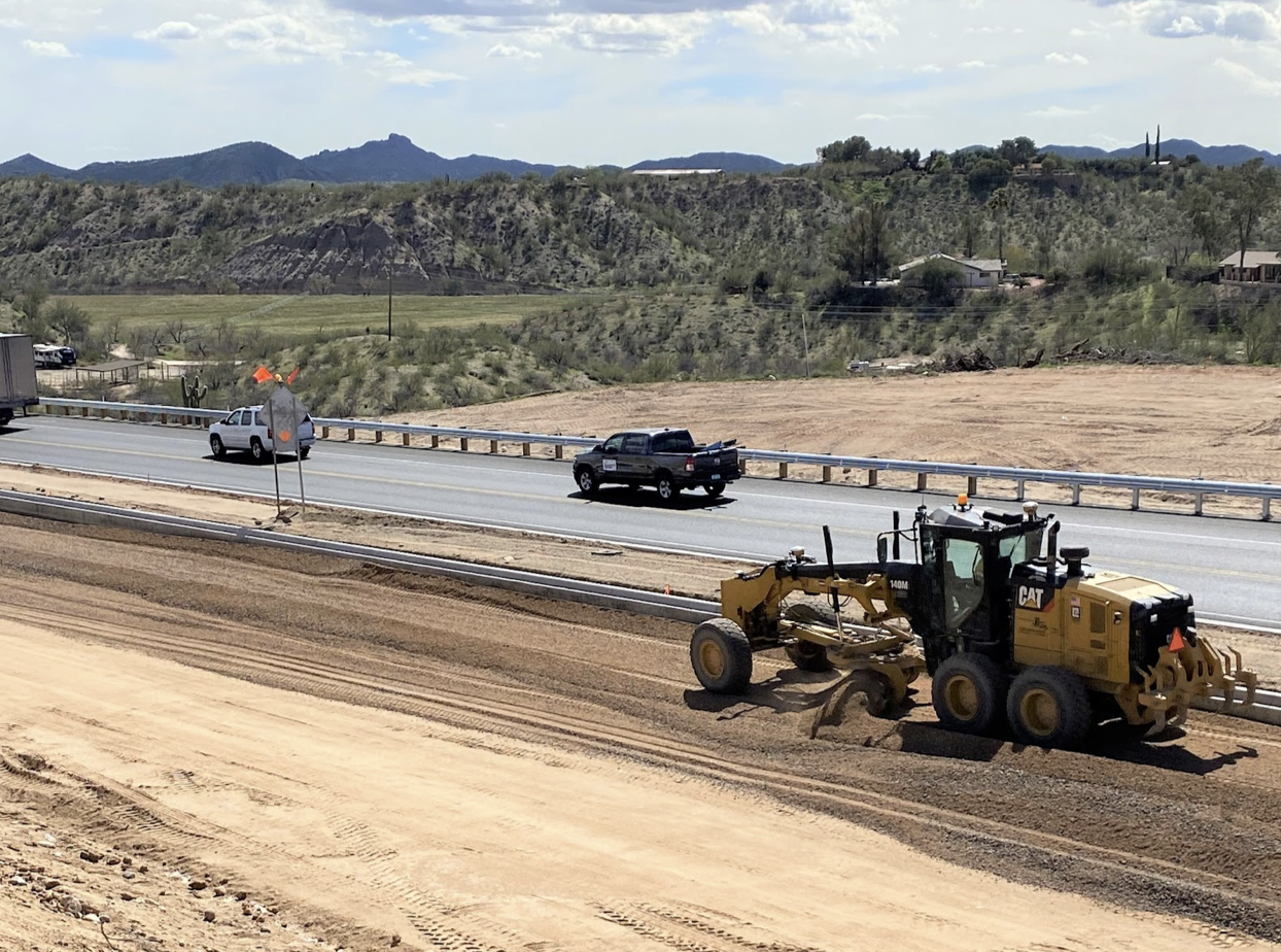 Heavy equipment works in a highway construction area as other vehicles drive by.