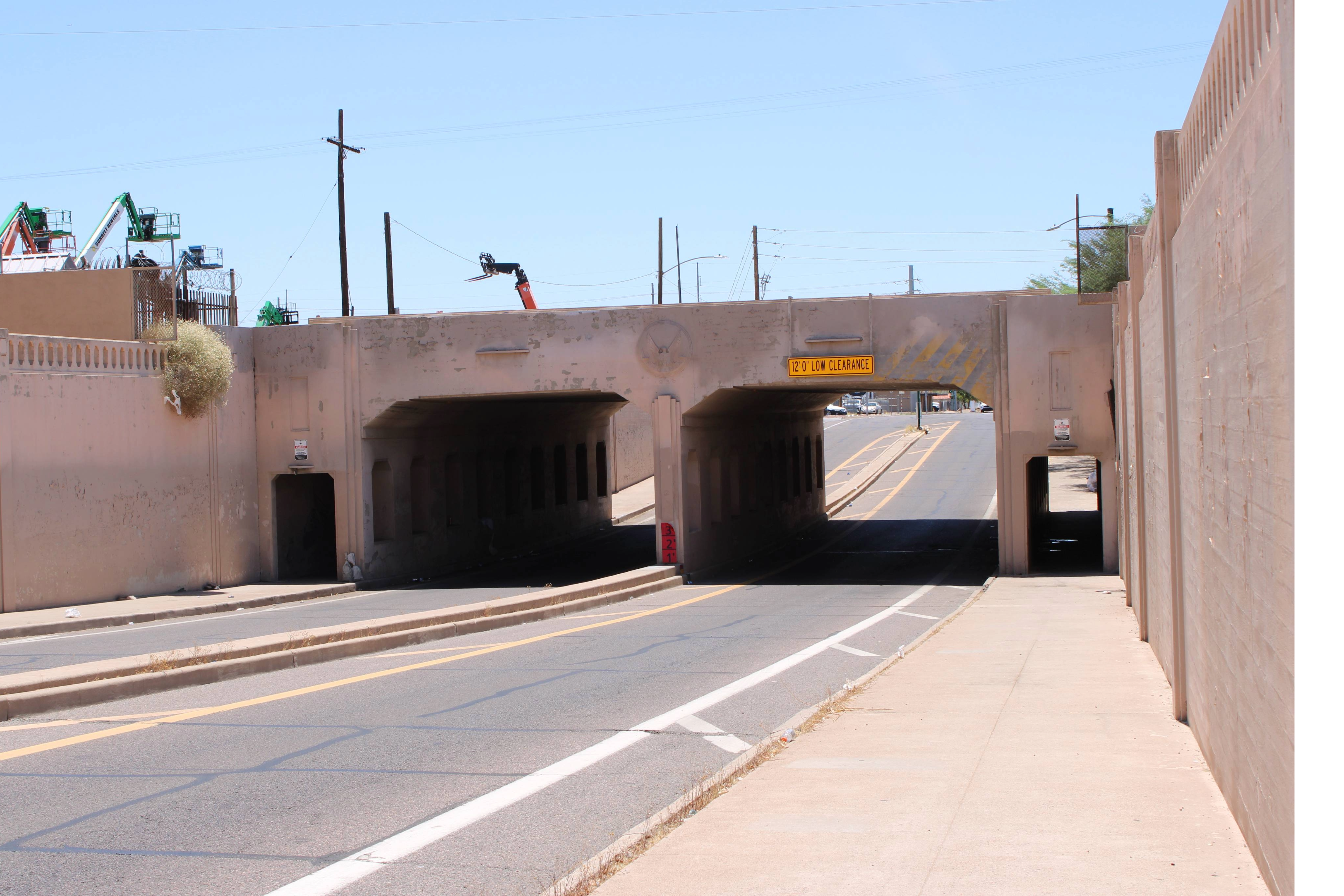A roadway with two low-clearance underpasses, bordered by a sidewalk and a fence, stretches into the distance under a bright, clear sky.