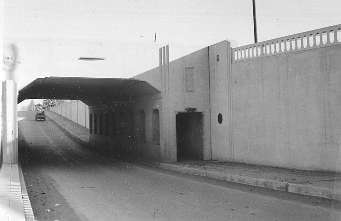A black and white photo of a car approaching an underpass beneath a bridge on a slightly inclined road. The sky is clear, and the surroundings are free of traffic and pedestrians.