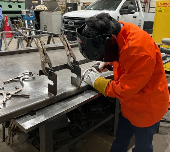 An individual in an orange safety jacket holding a welder and wearing a welding mask. 