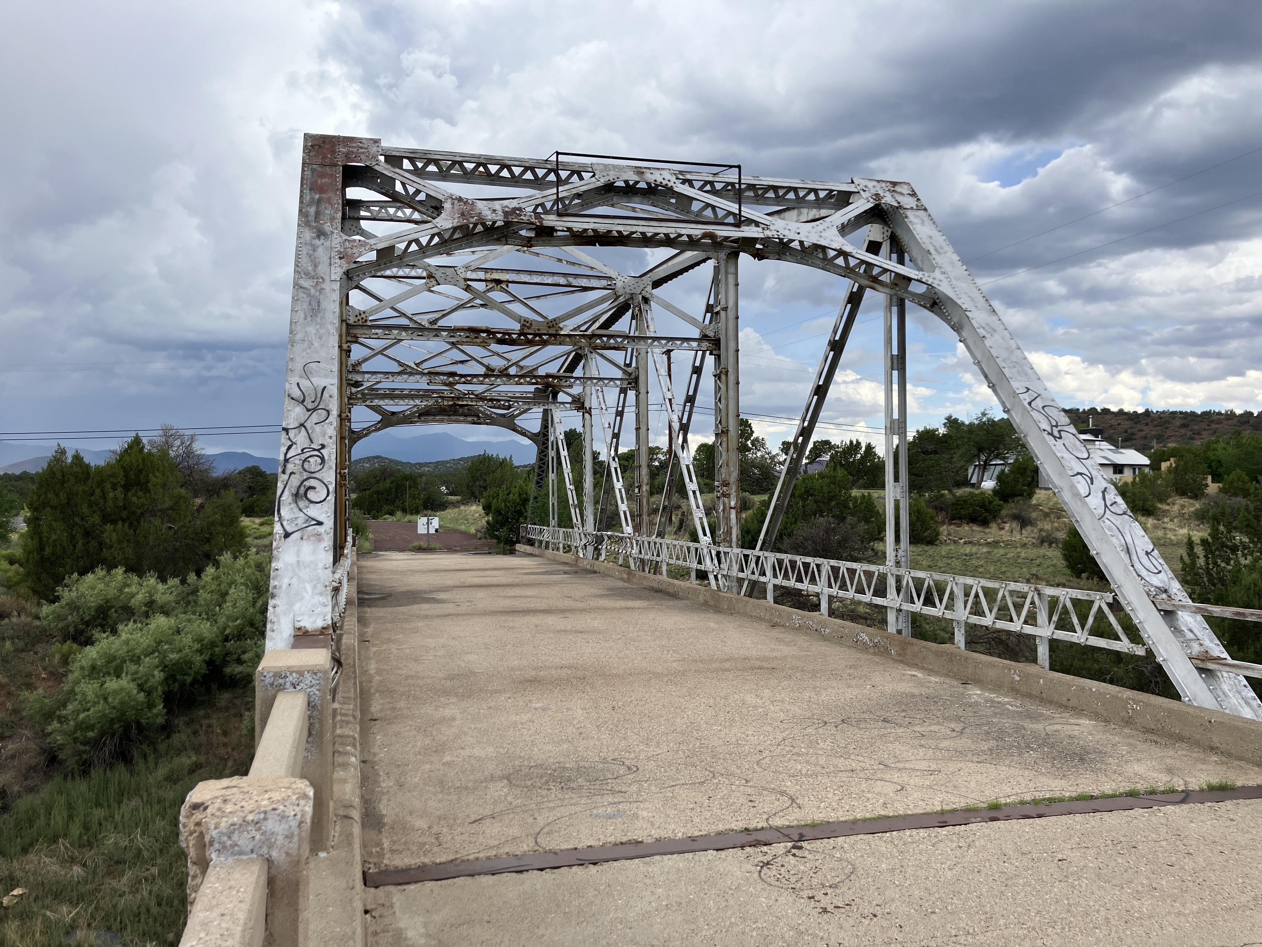 An old, weathered metal truss bridge with graffiti spans over greenery under a cloudy sky.