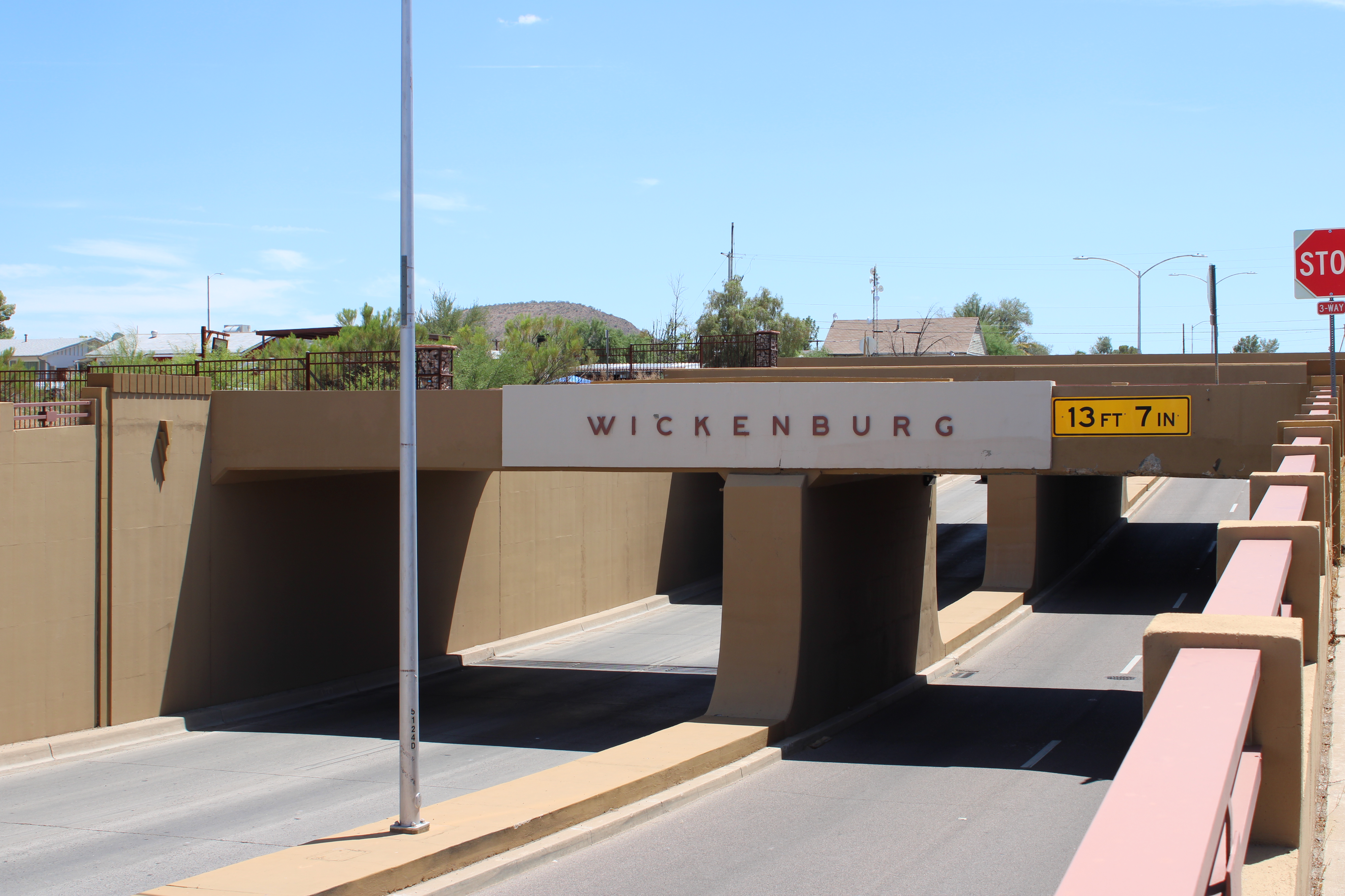 A road passes beneath a bridge with the name "Wickenburg" displayed on the side. A height restriction sign nearby indicates a clearance of 13 feet 7 inches.