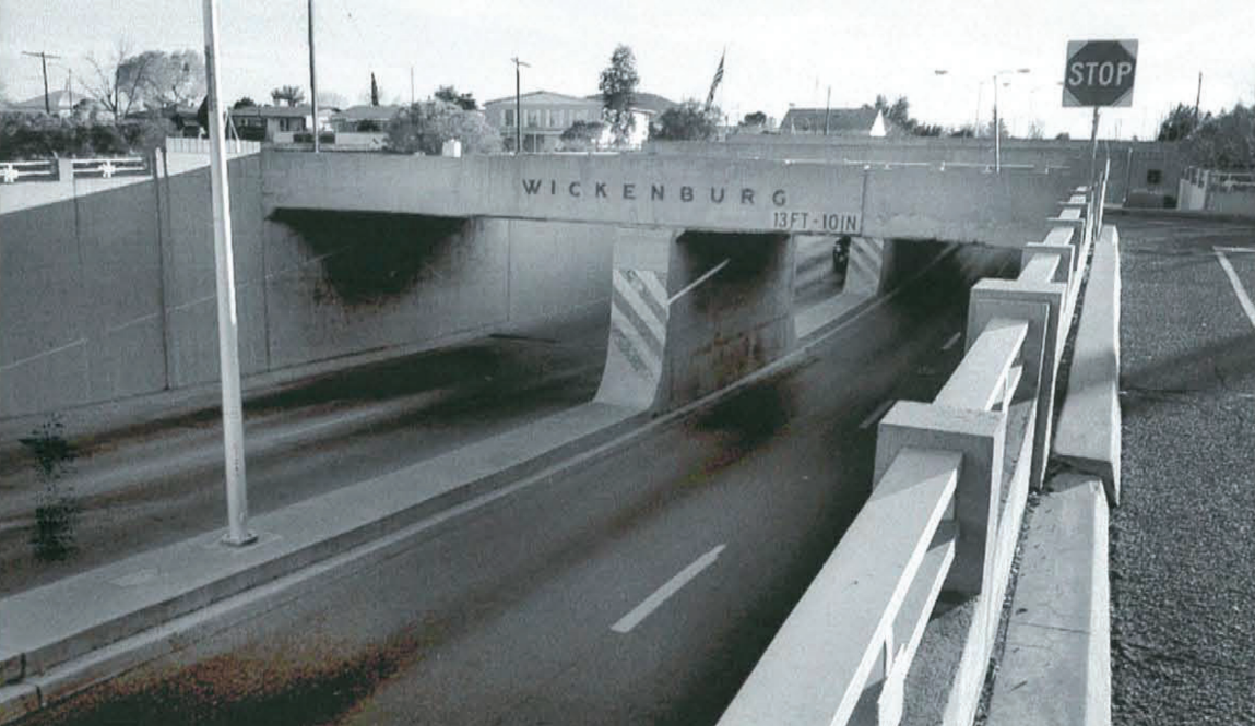 A concrete overpass with "WICKENBURG" and "1971-1971" written on it, spans across a road. There's a "STOP" sign visible on the right side.