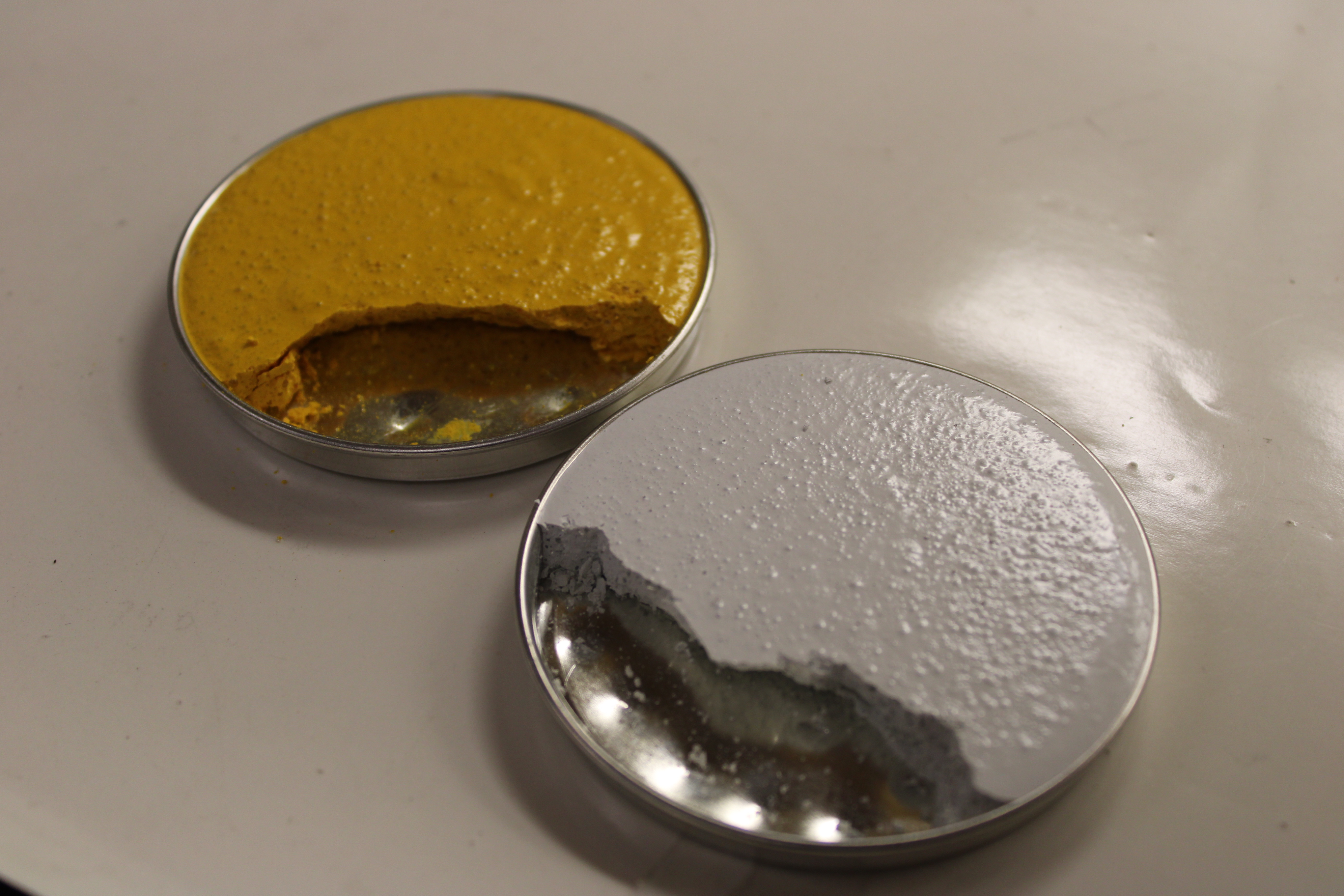 Two small metal tins filled with thick, textured substances: one with yellow paste and one with white paste. Both have a portion scooped out, showing the metal bottom of the tins.