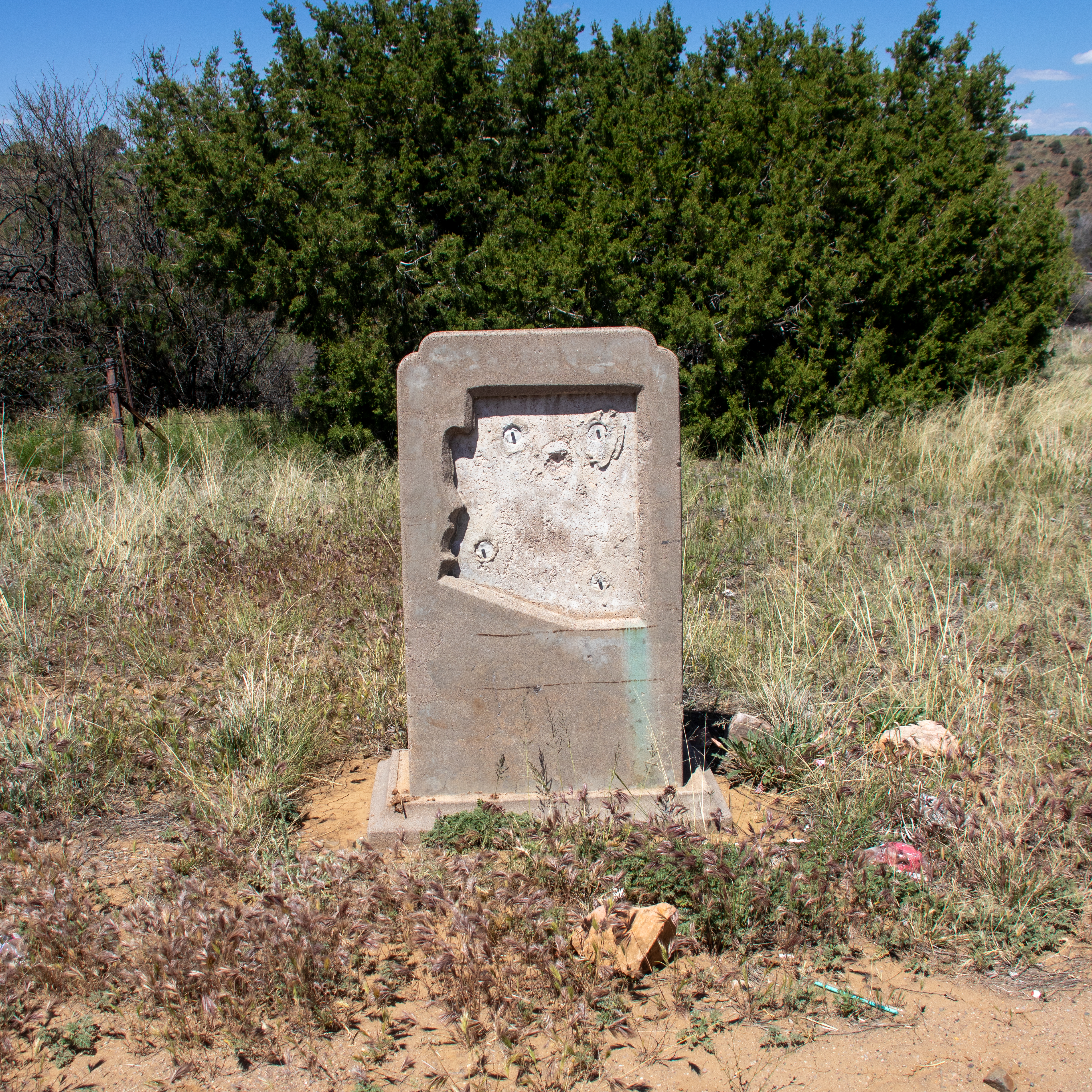 A stone marker along US 60 in Arizona is missing a metal plaque. The landscape is full of weeds, plants and a bush in the background.