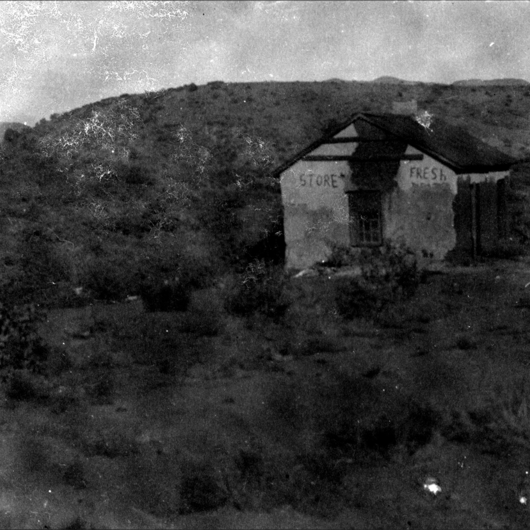 A dilapidated building in the ghost town of McMillanville Arizona with the faded words "STORE FRESH" stands on a hillside surrounded by rough terrain and shrubs. The town name was also spelled McMillenville.