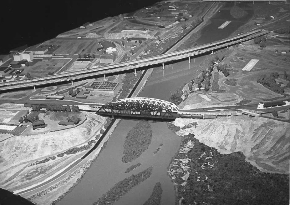 Model of a bridge and railway crossing over a river, with surrounding roads and landscape featuring buildings and fields.