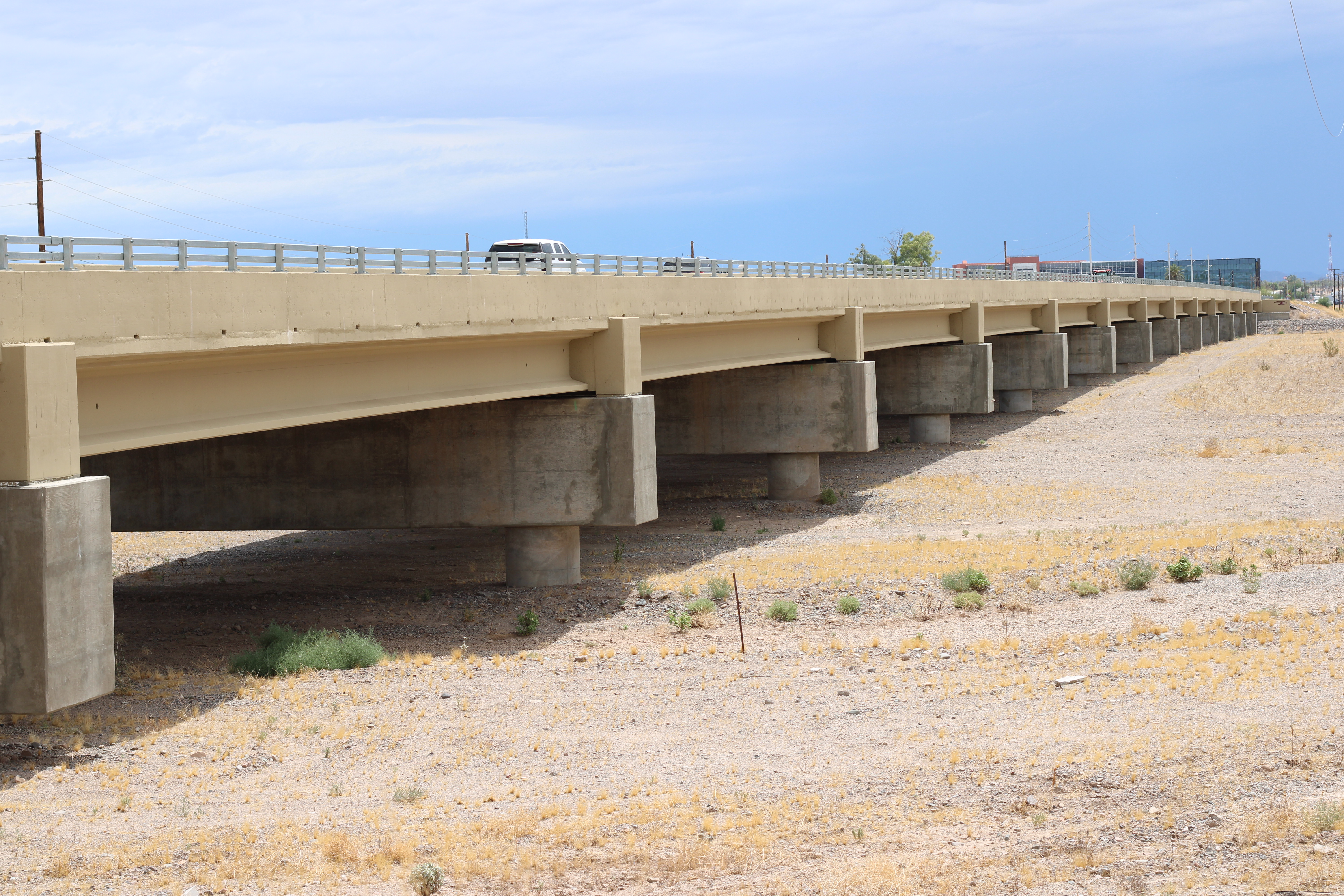 A concrete highway bridge spans a dry, sandy landscape under a cloudy sky. A vehicle is seen traveling on the bridge.