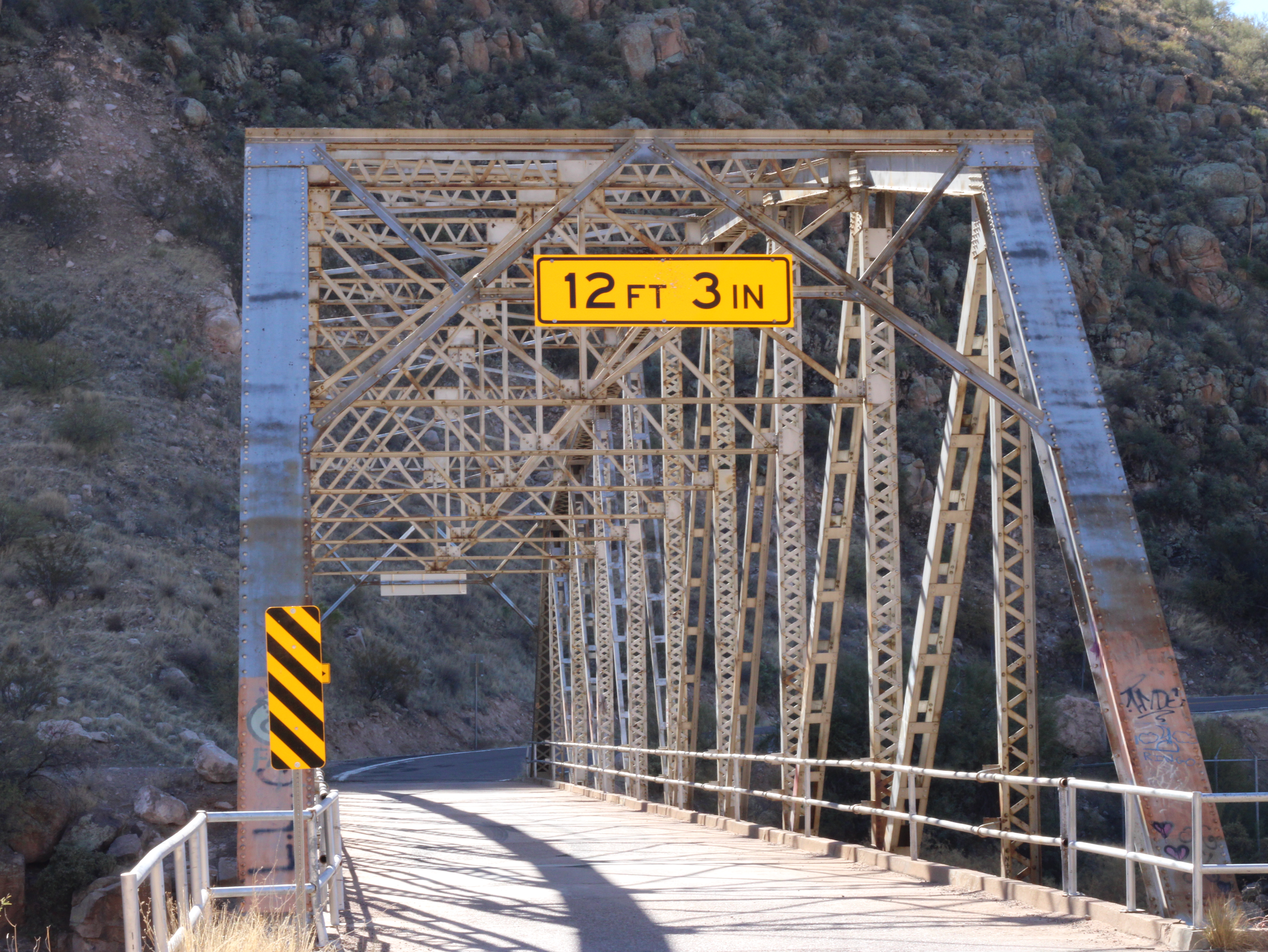 A steel truss bridge with a yellow clearance sign indicating 12 feet 3 inches. Rocky hillside visible in the background.