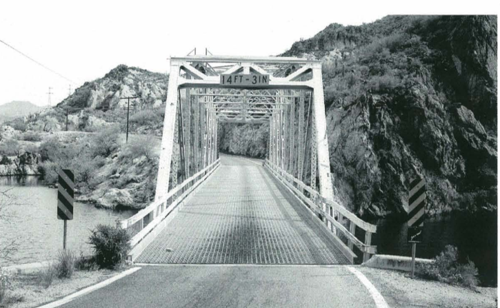 Metal truss bridge over a river, flanked by rocky hills, marked with a clearance sign of "14FT-3IN." Road signs and guardrails are present.