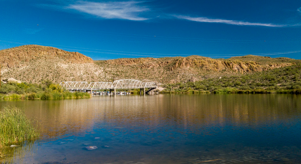 A metal truss bridge spans a calm river, surrounded by rocky hills, sparse greenery, and a blue sky with thin clouds.