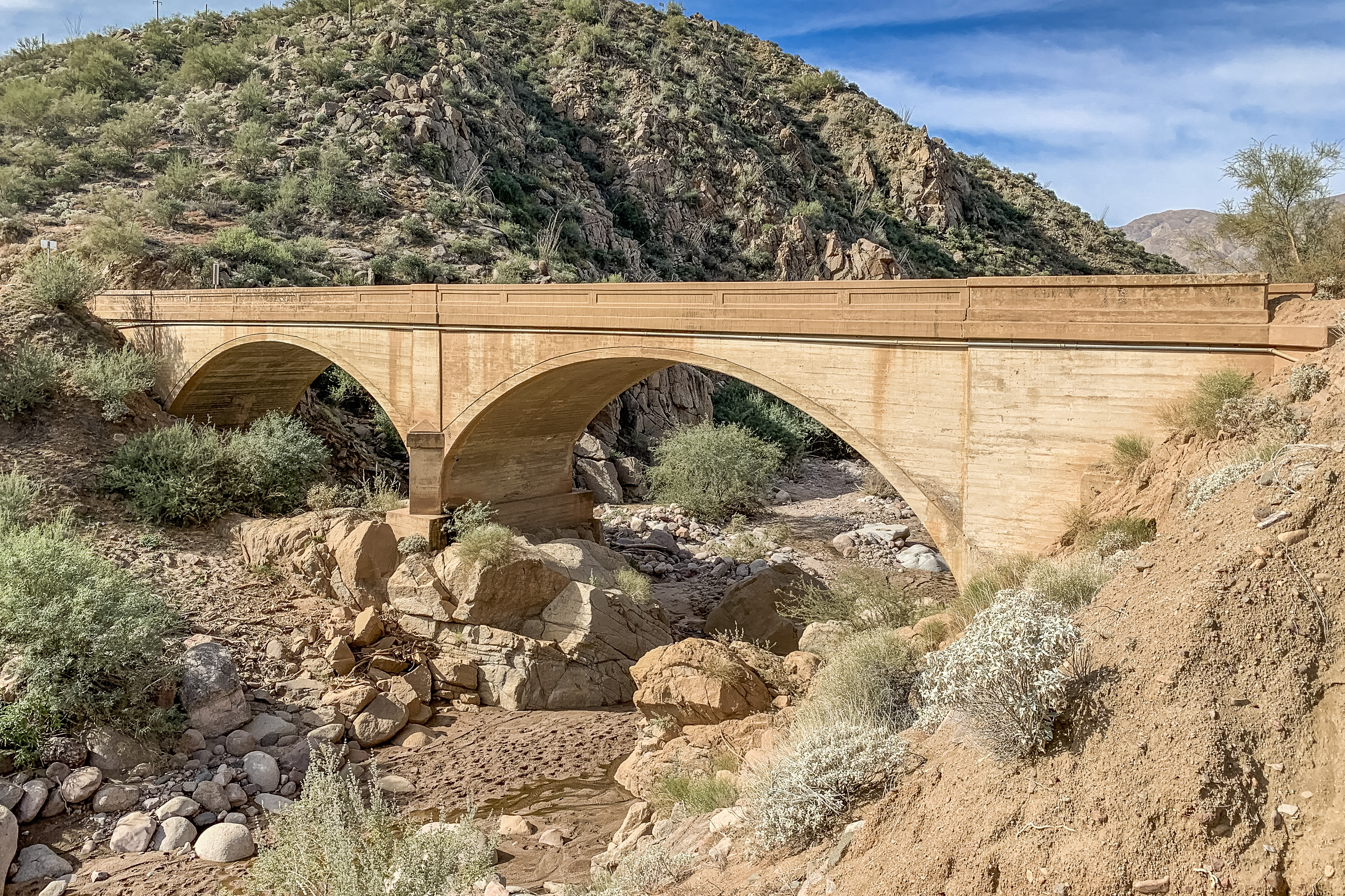A concrete arch bridge spans a dry, rocky creek bed surrounded by desert vegetation and hills.
