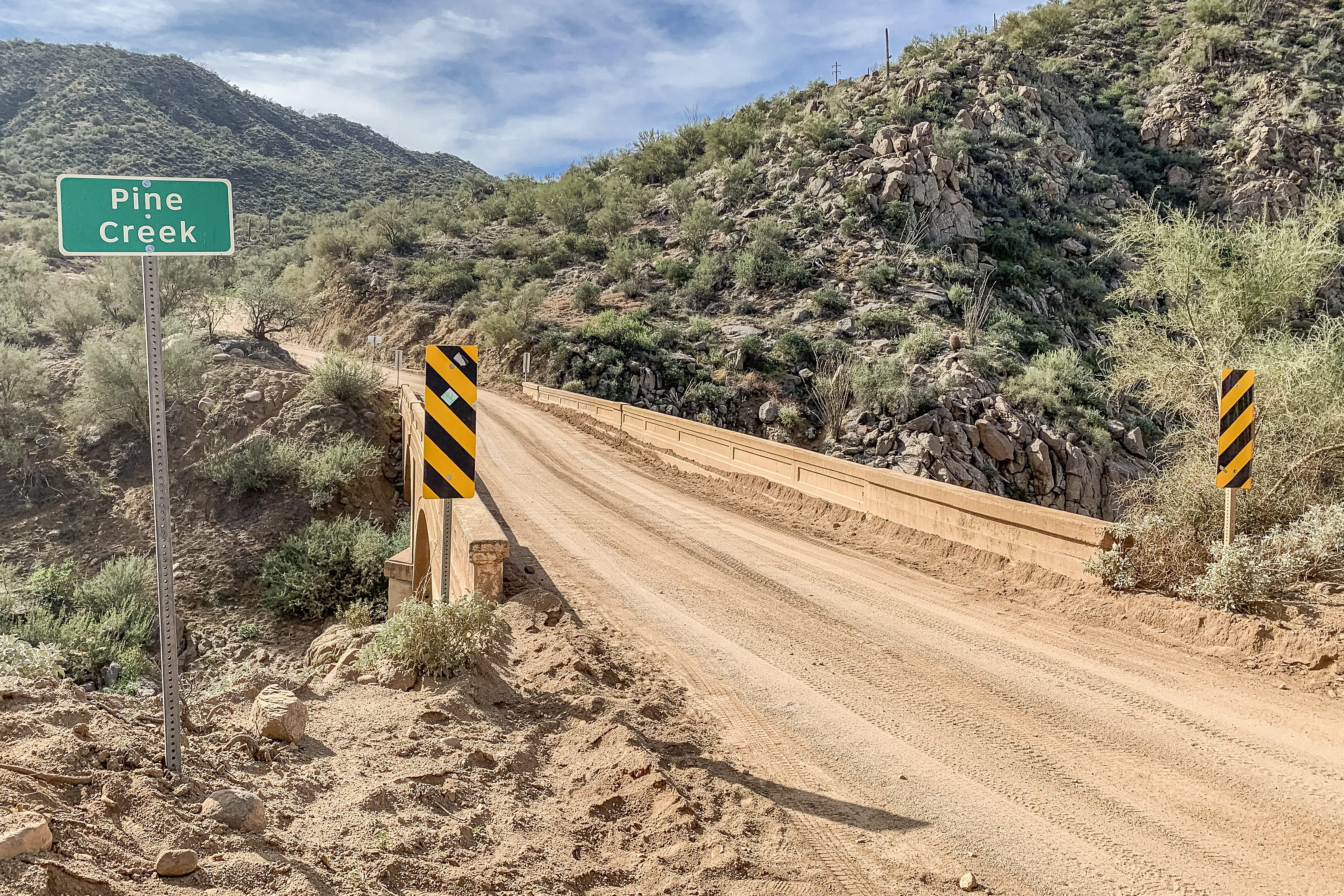 A dirt road leads to a small bridge over a dry creek bed, with a green sign reading "Pine Creek" and hills covered in desert vegetation in the background.
