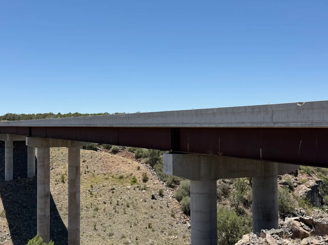 A concrete bridge with large support pillars spans over a dry, rocky landscape under a clear blue sky.