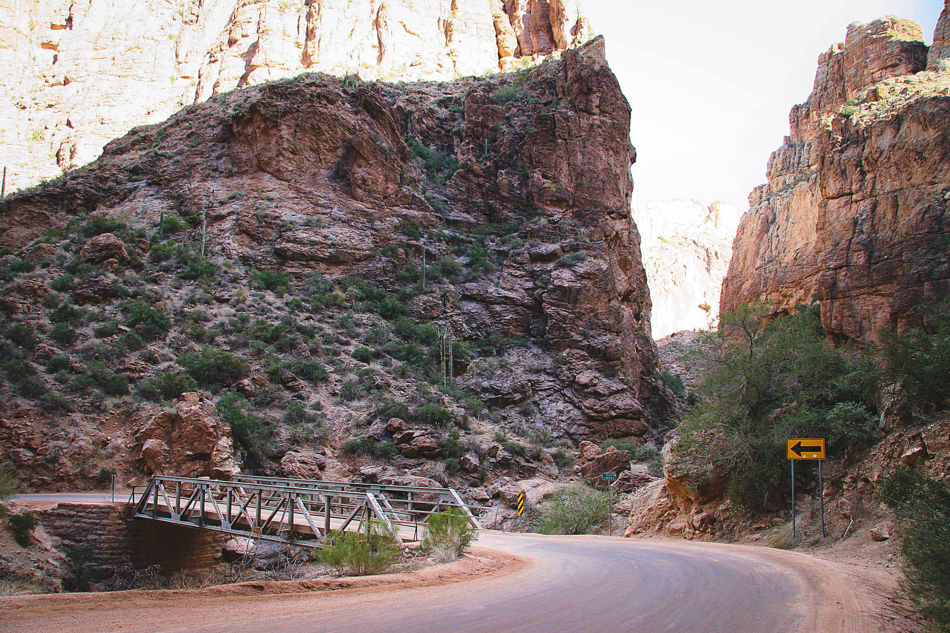 A metal footbridge spans a rocky canyon with steep cliffs and sparse vegetation under a bright sky.