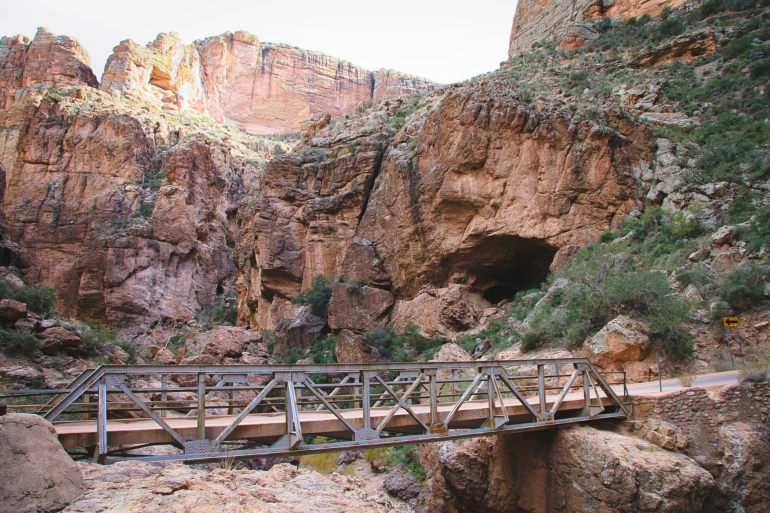 A metal footbridge spans a rocky canyon with steep cliffs and sparse vegetation under a bright sky.