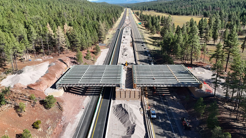 Aerial view of the I-17 Willard Springs Wildlife Overpass under construction, with cranes, trucks, and construction equipment on the highway surrounded by dense forest and mountains in the distance.
