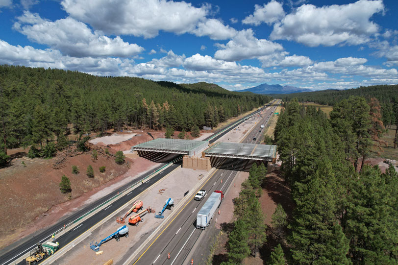 Aerial view of the I-17 Willard Springs Wildlife Overpass under construction, showing steel beams spanning across the highway surrounded by forest.