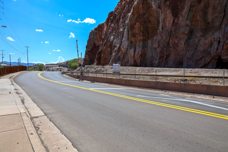 Paved section of US 191 curving alongside a tall rock face with fresh lane striping, railroad tracks, and utility poles under a bright desert sky.