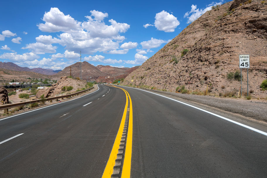 Paved section of US 191 with fresh lane striping, winding through desert hills with a 45 mph speed limit sign and a small town visible in the distance.