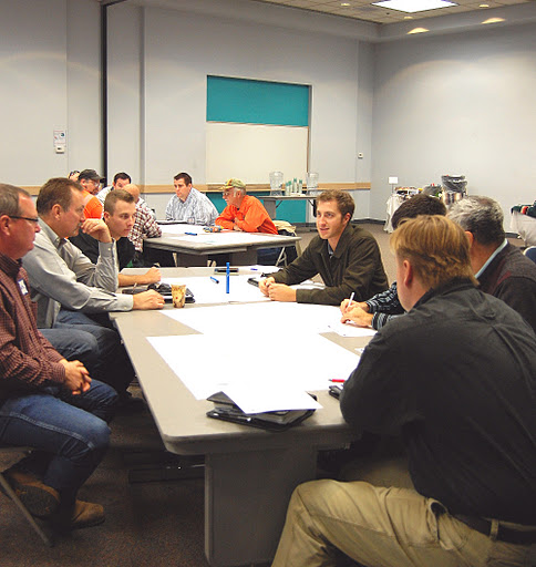 A group of people sit around tables in a meeting room, engaged in discussion and writing on paper.