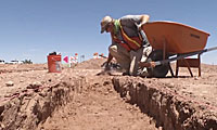 Person wearing safety gear excavates a trench at an archaeological site under a clear sky, with a wheelbarrow and orange bucket nearby.