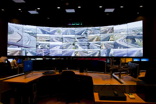 A control room with large display screens showing multiple live surveillance camera feeds of highways and roads, surrounded by workstations and computer monitors.