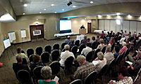 A speaker presents to an audience seated in a conference room with a projection screen at the front.