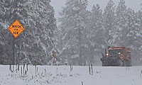 Snow plow on a snowy road with watch for ice sign