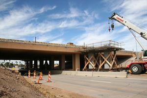 A crane and construction scaffolding are positioned near an overpass under repair, with traffic cones blocking part of the road.