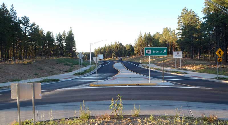 A newly paved road with a roundabout, surrounded by trees. A green highway sign indicates the direction to Sedona via State Route 89A.