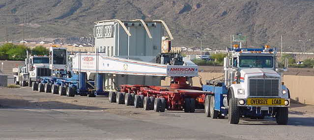 Three large trucks transport an oversized load on a multi-axle trailer along a road near mountains.