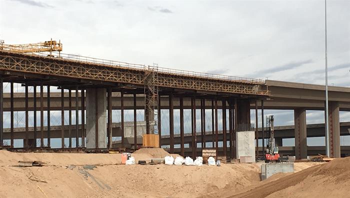A highway overpass under construction with scaffolding, cranes, and sand piles in the foreground.