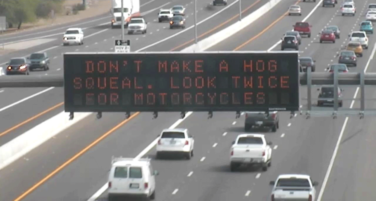 Overhead highway sign reads, Dont make a hog squeal. Look twice for motorcycles, above lanes of moving traffic.