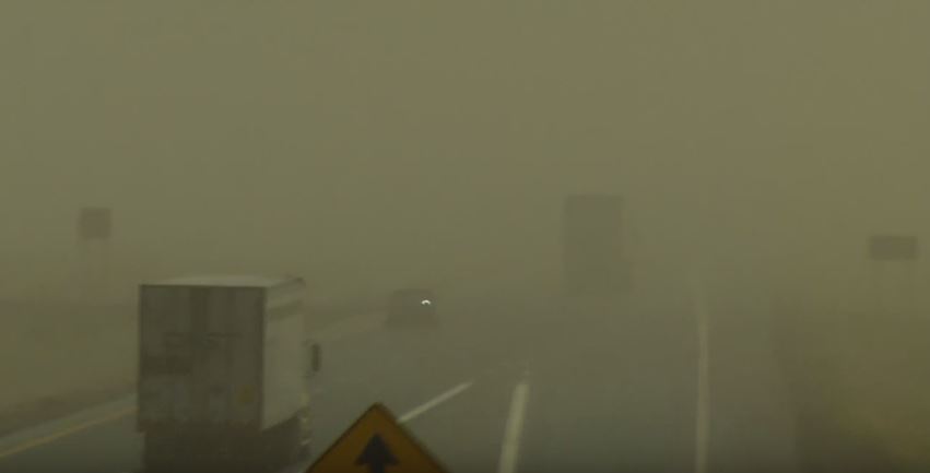 Trucks and vehicles drive on a highway with very low visibility due to heavy dust making road signs and surroundings barely visible.