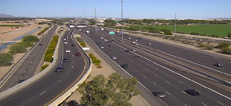 A wide freeway with moderate traffic flows beside fields and trees under a clear blue sky.