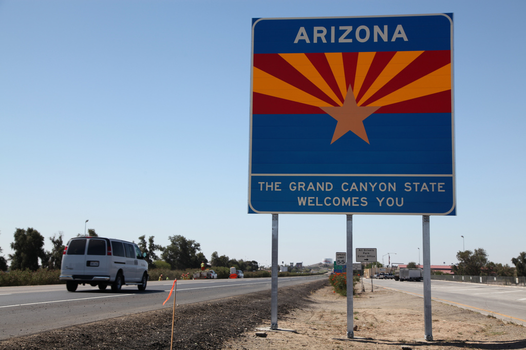A blue Arizona welcome sign by a highway reads The Grand Canyon State Welcomes You.