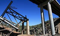 Collapsed bridge with twisted metal beams and concrete pillars under a clear blue sky.