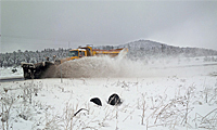 A snow plow clears snow from a road in a snowy landscape, spraying snow to the side. Snow-covered grass and hills are visible in the background under a cloudy sky.