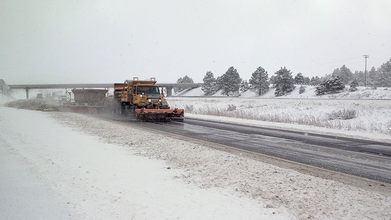 A snowplow clears snow from a highway on a snowy day, with trees and an overpass visible in the background.