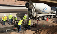 Construction workers in safety vests pour and level concrete from a cement truck at a roadside construction site.