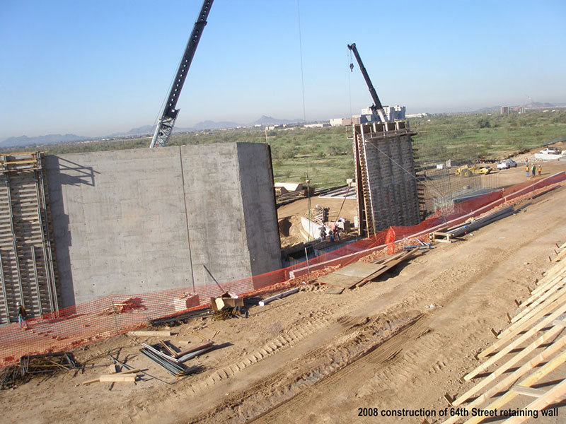 Construction site with cranes and workers building a large retaining wall, surrounded by dirt, materials, and safety barriers.
