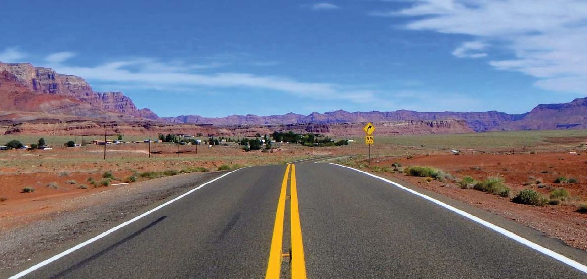 A two-lane highway with a double yellow line runs through a desert landscape with red rock formations under a blue sky. A road sign and small buildings are visible in the distance.
