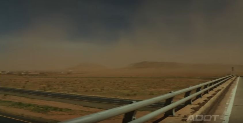 Dust storm approaching a desert highway, with guardrails and distant hills under a dark sky.